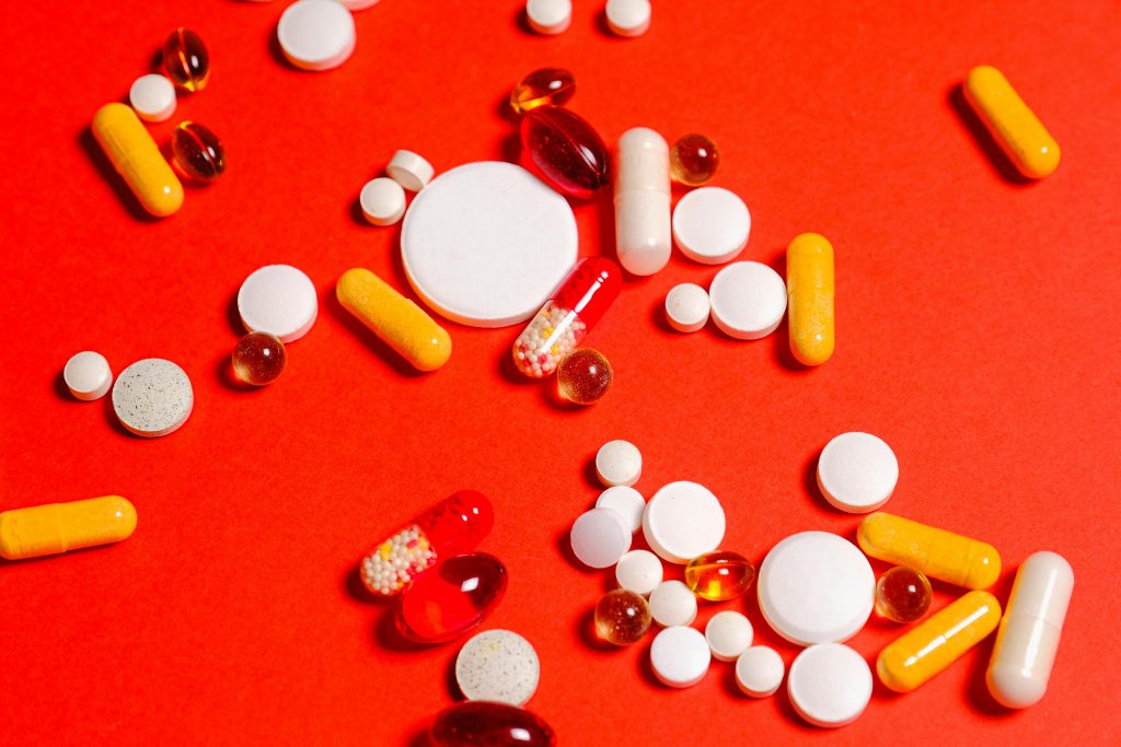 Variety of pills and capsules scattered on vibrant red backdrop, showcasing diverse medicine forms.